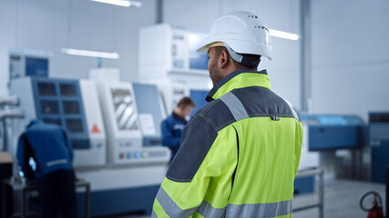 Industry 4 Factory: Modern Worker Wearing Safety Jacket and Hard Hat, Walks Through Contemporary Industrial Workshop where Professionals are Working and Programming CNC Machinery.
