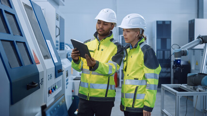 Chief Engineer and Project Manager Wearing Safety Vests and Hard Hats, Use Digital Tablet Computer in Modern Factory, Talking, Optimizing CNC Machinery, Programming Machine for Increasing Efficiency