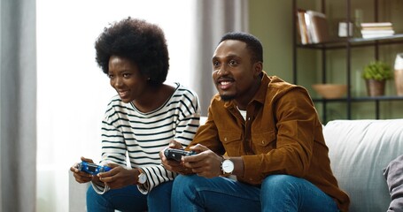 Young happy african american couple sitting at home on sofa and playing video games on console. Cheerful woman having fun winning over man in game.