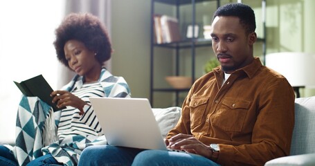 Young married african american couple spending leisure time together sitting on sofa at home. Man looking at laptop, woman reading book.
