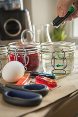 Frau gießt flüssige Ostereier Kaltfarbe Grün in Glas mit Essig Wasser auf Papier Unterlage in Küche neben Orange, Gelb, Blau, Rot in Verpackung, gekochte Eier und Schere zum Färben zu Ostern