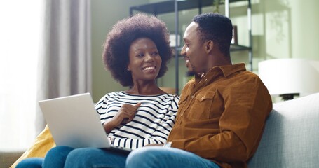 Young African American couple sitting at home on couch and talking to friends over video call using laptop.