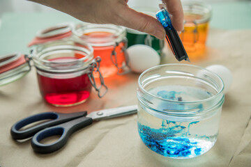 Frau gießt flüssige Ostereier Kaltfarbe Blau  in Glas mit Essig Wasser auf Pastell Papier Unterlage neben Orange, Grün, Gelb, Rot in Verpackung, gekochte Eier und Schere zum Färben zu Ostern