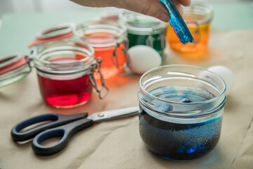 Frau gießt flüssige Ostereier Kaltfarbe Blau  in Glas mit Essig Wasser auf Pastell Papier Unterlage neben Orange, Grün, Gelb, Rot in Verpackung, gekochte Eier und Schere zum Färben zu Ostern