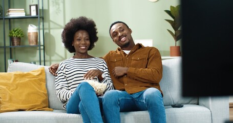 Young happy african american couple sitting at home on sofa and watching movie on tv and eating popcorn.