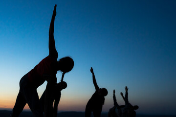 A group of silhouettes of people doing stretching and toning exercises after the running session or during a yoga class somewhere outdoors at sunset.