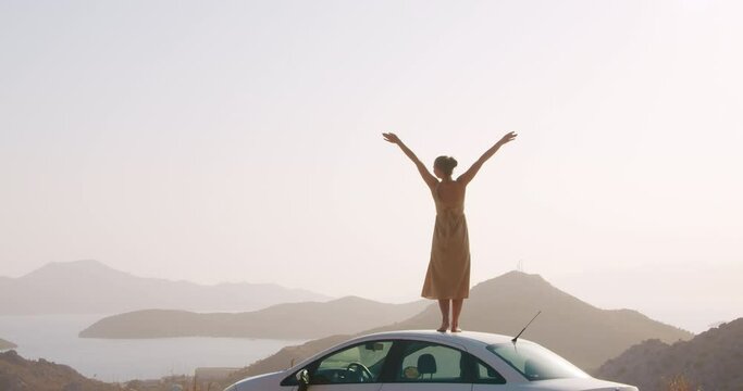 Young woman staying on car roof on road side on top of mountain and raises arms into air, happy, youth and happiness in fornt of sea landscape. Free as a bird concept. Road trip adventure
