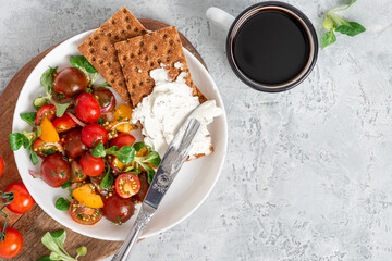 Tasty breakfast or lunch. Salad of colorful cherry tomatoes, onions, herbs, and olive oil, served with crispbread and cream cheese on a gray background top view. Free space for text.