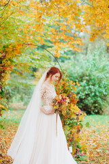 Obraz na płótnie Canvas Happy red-haired bride stands in autumn park with a bouquet of flowers in her hands.