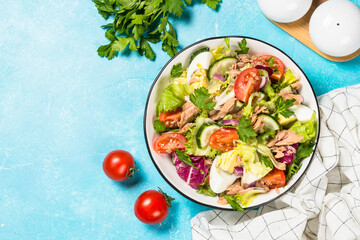 Tuna salad with green leaves, eggs and vegetables in white bowl. Top view at blue background.