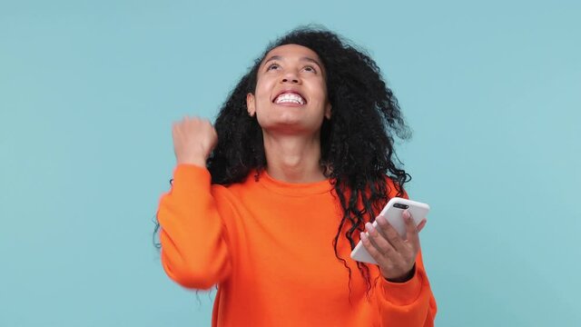 Happy fun young african american woman in orange sweatshirt hold using mobile cell phone typing browsing say wow yes just found out great big win news doing winner gesture isolated on blue background