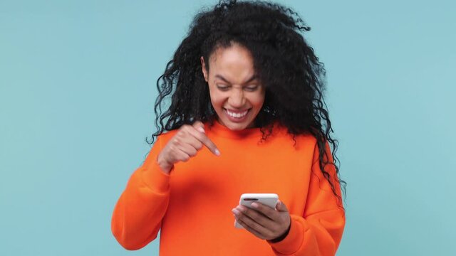 Shocked amazed fun young african woman in orange sweatshirt hold using mobile cell phone typing browsing say wow yes just found out great big win news doing winner gesture isolated on blue background