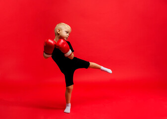 Fototapeta na wymiar a little boy boxer stands in boxing gloves and makes a lunge with his foot on a red background with a place for text