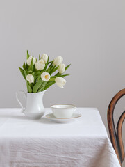 A bouquet of white tulips and a white cup on a white table and vintage chair beside. Easter table decor