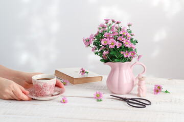 Woman's hands holding a vintage cup of coffee and pink chrysanthemum flowers in a pink vintage pitcher. Sunny table setting at home