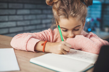 Close up photo of a blonde little girl drawing something in her notebook sitting at the table in the kitchen