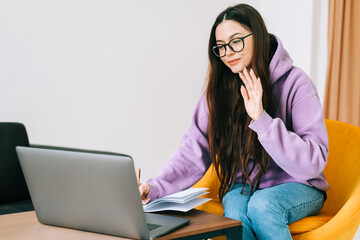 Young caucasian woman college student in glasses studying with books, laptop distantly at home using video call, weaving. Distantly education concept.