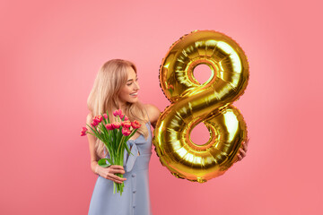 Sexy young lady with flowers and golden balloon shaped as number eight over pink studio background