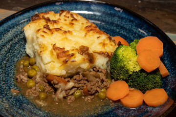 A delicious plate of Shepherds Pie with Mash Potato, Carrots and Broccoli on a wooden kitchen table