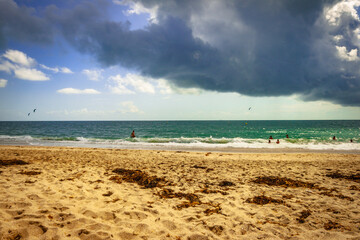 Plage de sable avec des algues sur le sable et des personnes dans l océan au dessu un ciel menaçant avec des nuages gris noirs .