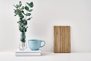 A mug on a stack of books and a transparent vase with eucalyptus branches, a wooden board against the wall. Eco-friendly materials in interior decor, minimalism. Copy space, mock up.