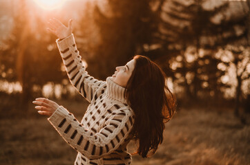 Warm portrait of a young woman wearing a comfy sweater waving arms at sunset light with trees and lake in the background