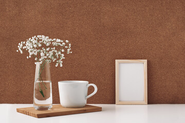 Frame, vase with white flowers and a cup, cork background. Mock up, copy space. Folk.