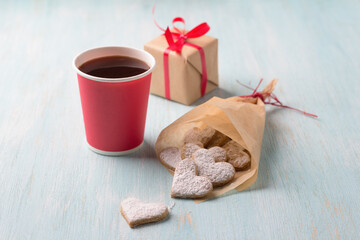 Delicious homemade heart shaped cookies sprinkled with powdered sugar in craft bag for valentine's day gift with paper cup of coffee, on light blue background