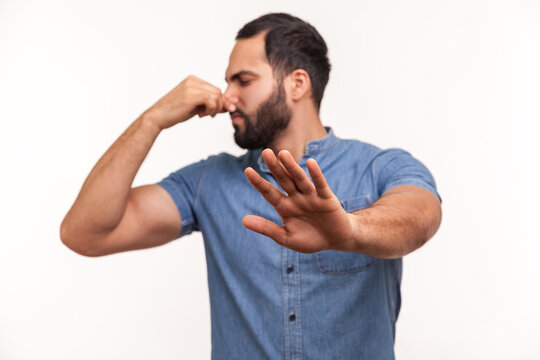 Confused unhappy bearded man grabbing nose with fingers and stretching hand out feeling disgusted unpleasant smell