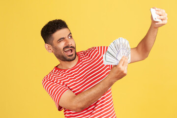 Extremely happy satisfied man blogger in striped t-shirt making selfie photo with bunch of dollar...