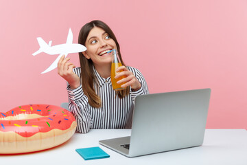 Positive woman drinking lemonade holding paper plane in hand, planning vacation abroad sitting at workplace with laptop, passport and rubber ring on desk
