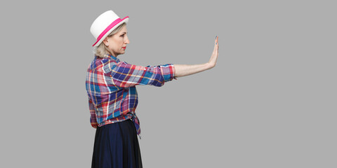 Stop it. Profile side view portrait of serious modern stylish mature woman in casual style with hat standing looking forward and showing stop sign.