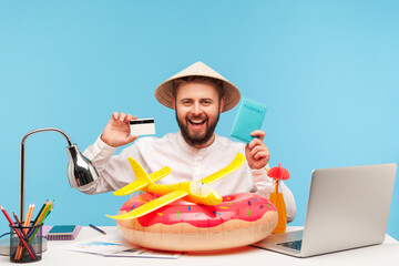 Happy positive man sitting at workplace with rubber ring and toy plane, holding passport and credit...