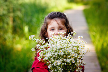 Selective image focus. A small long-haired girl in a burgundy dress stands on a wooden bridge with a bouquet of daisies. She plays with the bouquet in front of her.