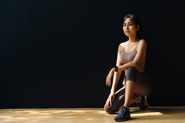 Focused young Asian sportswoman relaxing and getting ready for workout while crouching in studio on black background - full body length