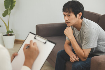 Young asian man, male has mental symptoms which must have been therapy and stress, sitting on couch to consult to psychologist during the session  taking notes to find out how to treat the therapist.