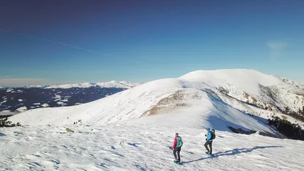Fotobehang Een drone-opname van een stel dat sneeuwschoenen draagt en naar de top van de Speikkogel in de Oostenrijkse Alpen wandelt. De hele helling is bedekt met sneeuw. veel bergketens in de rug. Winterse buitenactiviteit. Plezier © Chris