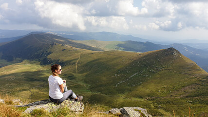 Fototapeta premium Woman in hiking outfit sitting at a big rock and enjoys the view, while hiking to Speikkogel, Austrian Alps. The mountains around are lush green, not so sharp. Sunny summer day. Serenity and calmness