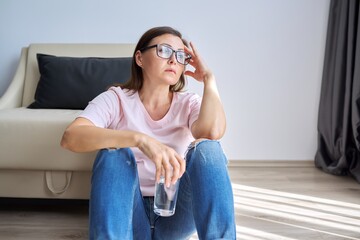 Sad unhappy mature woman sitting at home on floor with glass of water