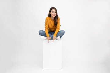 Smiling young Asian woman sitting on white box and pointing down empty copy space isolated on white background - 408353903