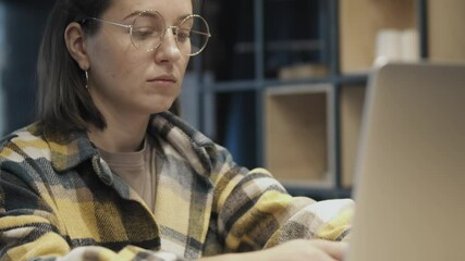 Close-up of young casual woman in round glasses sitting at desk at home and browsing laptop relaxing alone.
