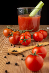 Top view of cherry tomatoes with peppercorns, wooden spoon, glass of tomato juice with celery, selective focus, on wooden table, black background, vertical