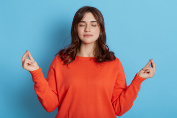 Young good looking woman meditates, keeps both hands in okay gesture, and stands with closed eyes,...
