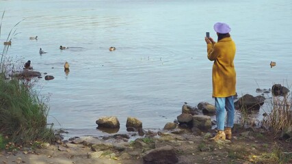 woman in autumn clothes taking photo on smartphone near lake with ducks