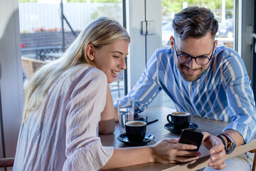 Young romantic couple enjoying a cup of coffee together in a cafe, sitting at the table and talking while scrolling through social media on the smartphone.