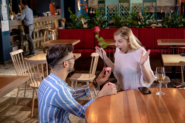 Young romantic couple on a date in a restaurant. Boyfriend making proposal to his girlfriend with a red rose and an engagement ring on valentine day.