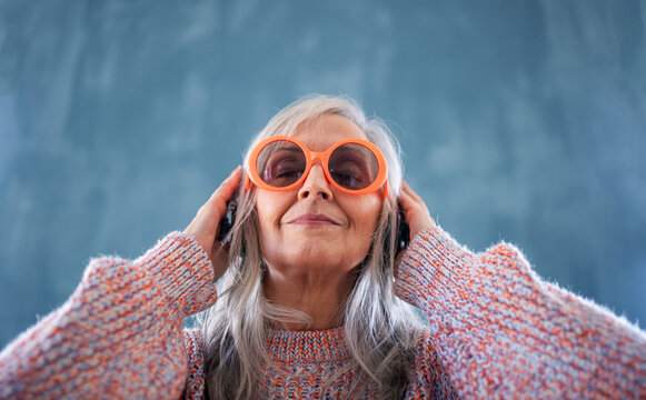 Portrait Of Senior Woman With Sunglasses Standing Indoors Against Dark Background, Listening To Music.