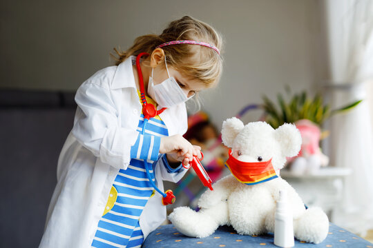 Little girl makes injection to teddy bear. Cute child with medical mask playing doctor, holding syringe with vaccine. Coronavirus covid vaccination concept. Kid play role game at home.