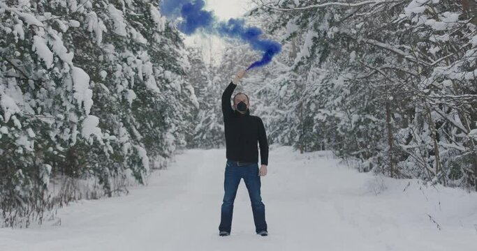 Man in forest with blue smoke grenade, bomb. A masked man with a blue smoke bomb in a white clean forest. Concept - freedom, protest, signal for help, ecological catastrophe. Slow motion