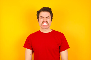 Fototapeta premium young Caucasian man wearing red t-shirt standing against yellow background keeps teeth clenched, frowns face in dissatisfaction, irritated because of much duties.
